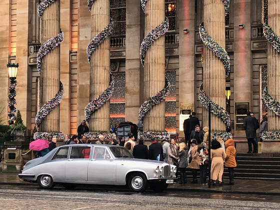 Christmas decorations wrapped around columns outside a building, silver Rolls Royce, and a crowd of people.