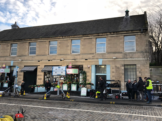 Film crew and crane outside cafe with blue window frames, cobbled street.