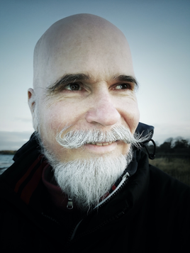 head shot of man with bald head, dark eyebrows, white beard and moustache, outside on a cold day with sea behind him.