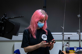 young woman with long pink hair in a black t-shirt wearing a blue paper face mask.