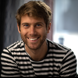 Smiling young man with short hair and facial stubble wearing a black and white stripy stop.