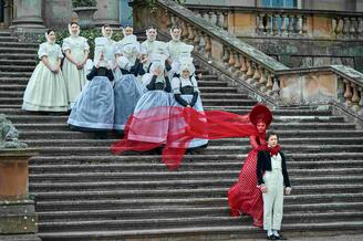 woman in red dress and veil holding on to boy in historic clothing, with girls in white frocks behind her, standing on wide steps of a mansion house.